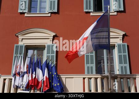 Rote, historische Gebäudefassade in Nizzas Altstadt mit flatternden französischen Trikolore-, Regional- und EU-Flaggen auf einem reich verzierten Balkon. Stockfoto
