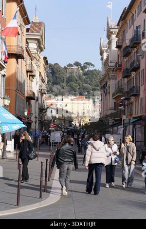 Lebhafte Fußgängerzone in Vieux Nice mit farbenfrohen Pastellfassaden, französischen Flaggen und Menschen, die in Richtung des grünen Hügels Colline du Château schlendern. Stockfoto