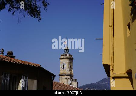 Historischer Glockenturm mit Flagge in der Altstadt von Vieux Nizza, eingerahmt von gelben Gebäuden unter klarem blauem Himmel. Nizza, Frankreich Stockfoto