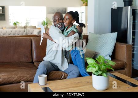 Der ältere afroamerikanische Großvater und die Kind-Enkelin umarmen sich auf einem Ledersofa mit Papier Stockfoto
