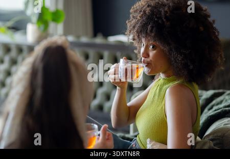 Verschiedene weibliche Freunde schlürfen aus klaren Glasbechern auf einem getufteten Sofa und tragen ärmelloses grünes Oberteil Stockfoto