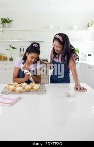 Afroamerikanische Mutter und Tochter dekorieren Cupcakes mit Gebäckbeutel in weißer Küche auf Schürzen Stockfoto