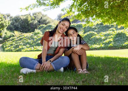 Afroamerikanische Mutter und Tochter sitzen auf Rasen unter Bäumen, tragen Rosthemd und Turnschuhe Stockfoto
