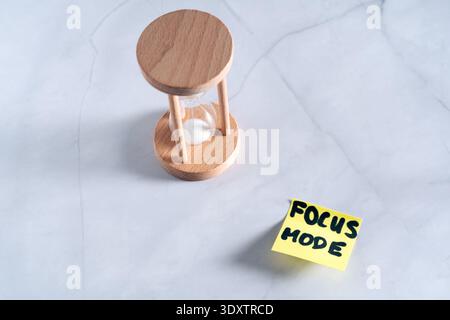Ein Bild über dem Kopf, der auf eine hölzerne Sanduhr blickt, und eine gelbe Post-it-Notiz mit FOKUSMODUS darauf, platziert auf einem Marmorhintergrund Stockfoto