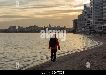 Eine Person spaziert am Sandstrand bei Sonnenuntergang in Torrevieja, Spanien, mit modernen Gebäuden am Ufer. Stockfoto