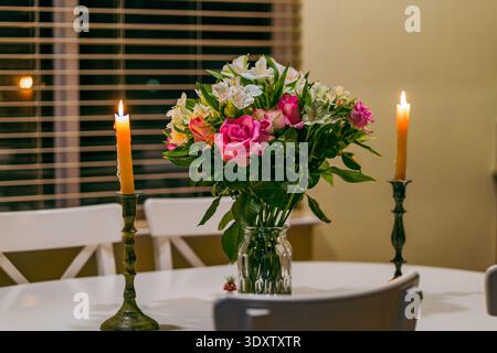 Ein leuchtender Strauß aus rosa Rosen und weißer Alstroemeria sitzt in einer Glasvase, flankiert von zwei leuchtenden orangefarbenen Kerzen auf einem weißen Esstisch. Stockfoto