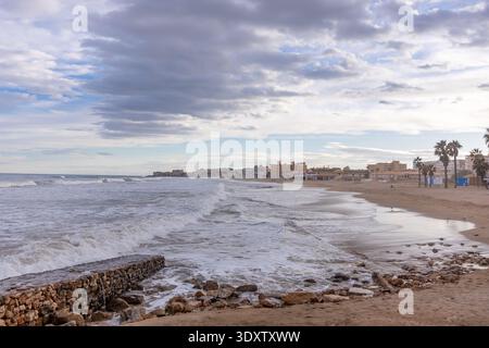 Eine Küstenszene in Torrevieja, Spanien, mit Wellen, die auf die Sandküste stürzen, einem Steinbruch und Palmen, die die Promenade unter einer bewölkten Oberfläche säumen Stockfoto