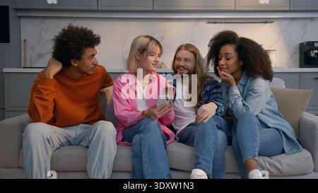 Multirassische Gruppe vier Freunde sitzen Couch zu Hause lächelnd lachen zusammen kaukasische Männer afroamerikanische Frauen diskutieren Nachrichten Handy Stockfoto
