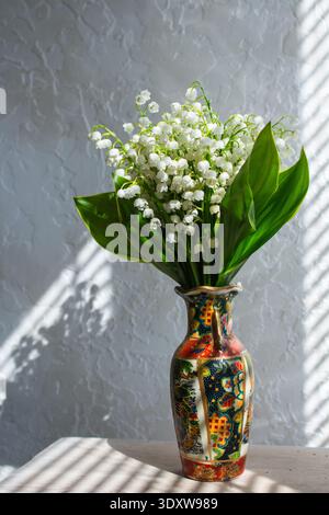 Frühlingsblumen Maiglöckchen aus dem Tal in dekorativer Vase. Sonnenlicht auf der Fensterbank. Weiße glockenförmige Blüten mit großen grünen Blättern Stockfoto