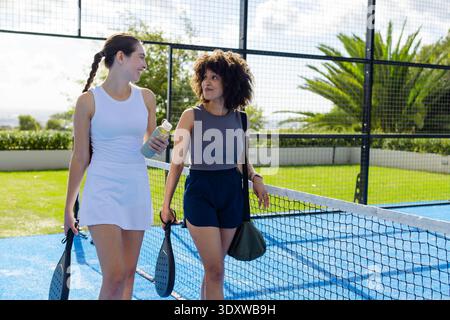Verschiedene weibliche Freunde in Turnkleidung, die auf dem blauen Padel-Platz laufen, Paddel halten, Wasserflasche Stockfoto