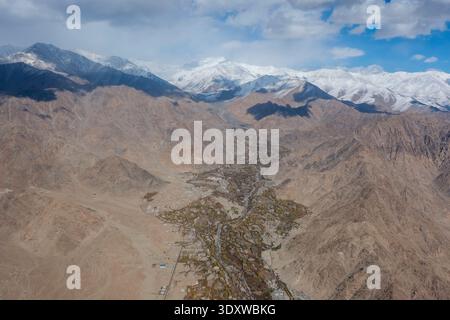 Aus der Vogelperspektive des oberen Teils von Leh mit einem städtischen Tal, eingebettet zwischen trockenen Bergen, mit der schneebedeckten Ladakh Range in der Ferne. Stockfoto