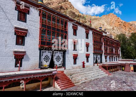 Ein buddhistischer Mönch in roten Gewändern geht über den Steinhof, vorbei an den verzierten, dunklen Holzfenstern und Dharmachakra-Vorhängen von Hemis Gompa. Stockfoto