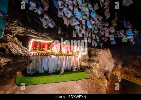 Banknoten sind an der niedrigen Felsendecke über einem beleuchteten Altar mit kleinen Statuen und rituellen Opfergaben in der Gotsang-Höhle in Hemis befestigt. Stockfoto