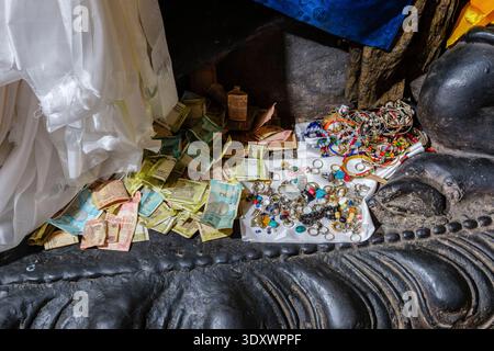 Schnäppchen der Indischen Rupie und verschiedene Schmuckstücke zu Füßen einer religiösen Statue auf einem dunklen Sockel in Thiksey Gompa. Stockfoto