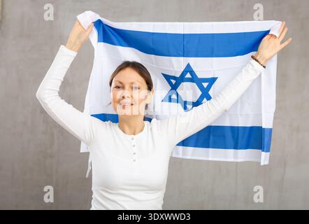 Positive patriotische Frau mittleren Alters, die die Staatsflagge Israels vor grauem Mauerhintergrund hält Stockfoto