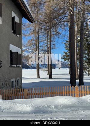Schneebedeckte Landschaft in Sils Maria, Engadin, Schweiz, mit Bauernhof, gefrorenem See und Berggipfeln unter klarem Winterhimmel. Stockfoto