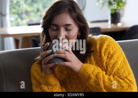 Frau in ihren 20ern in senfgelbem Strickpullover mit grauem Becher, das Aroma im Wohnzimmer einatmet Stockfoto