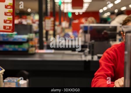 Verschwommenes Interieur im Supermarkt mit Kassierer in roter Uniform und Preisschildern. Professioneller Bokeh-Hintergrund, der den Einkauf eines Lebensmittelgeschäfts zeigt Stockfoto