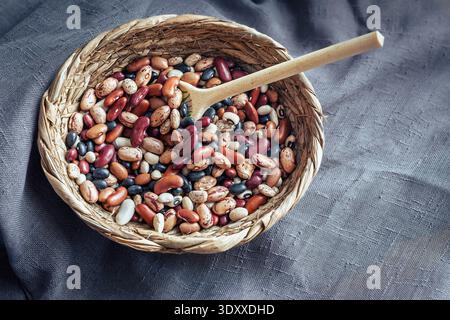Verschiedene Sorten getrockneter roher Bohnen und Hülsenfrüchte in einem gewebten Korb auf einem rustikalen dunklen Stoffhintergrund. Stockfoto