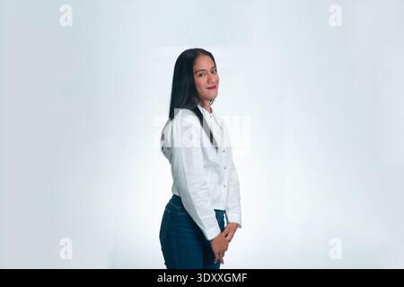 Ein einfaches Seitenporträt einer jungen indonesischen Frau, die ein weißes Hemd trägt und auf weißem Atelierhintergrund in die Kamera lächelt. Stockfoto