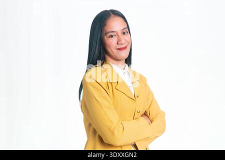 Porträt einer erfolgreichen indonesischen Frau in gelbem Blazer, die mit überkreuzten Armen und einem selbstbewussten Lächeln posiert, isoliert auf weißem Studiohintergrund. Stockfoto