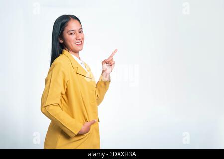 Fröhliche indonesische Frau in gelbem Blazer, die ihren Finger nach oben zeigt, während sie in die Kamera schaut, isoliert auf einem sauberen weißen Studiohintergrund. Stockfoto