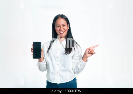 Porträt einer lächelnden indonesischen Frau in einem professionellen weißen Hemd, die ein Mobiltelefon mit einem leeren schwarzen Bildschirm hält und mit dem Finger auf die s zeigt Stockfoto