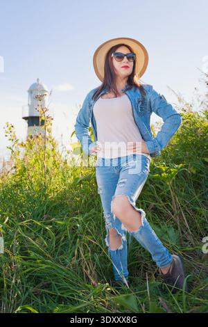 Flaches Porträt einer eleganten Frau in zerrissener Jeans, Jeansjacke und Strohhut. Das Modell steht selbstbewusst auf einem grünen Feld an einem sonnigen Tag, mit Stockfoto