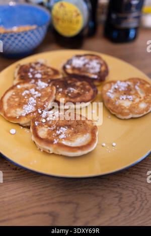Ein Teller Pfannkuchen mit Puderzucker drauf. Die Pfannkuchen sind kreisförmig auf einem gelben Teller und Holztisch angeordnet. Stockfoto