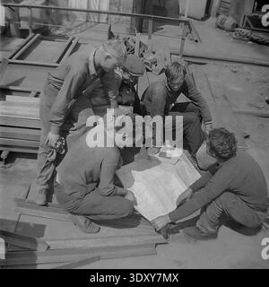 Unglaubliches historisches Schwarzweißbild, das eine Gruppe von fünf engagierten Fabrikarbeitern erfasst, die in den 1960er Jahren aktiv einen großen technischen Plan im Freien im Stroymash-Werk in Sloviansk analysierten Die geschickten Kameraden in lässiger Arbeitskleidung versammeln sich eng um den Papierplan und weisen im hellen Sonnenlicht auf mechanische Details hin. Dieser hochinteressante dokumentarische Schnappschuss zeigt meisterhaft sowjetische Ingenieursarbeit, kollektive intellektuelle Arbeit und die positive, konstruktive tägliche soziale Realität eines friedlichen Donbass vor dem Krieg. Stockfoto