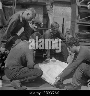 Unglaubliches historisches Schwarzweißbild, das eine Gruppe von fünf engagierten Fabrikarbeitern erfasst, die in den 1960er Jahren aktiv einen großen technischen Plan im Freien im Stroymash-Werk in Sloviansk analysierten Die geschickten Kameraden in lässiger Arbeitskleidung versammeln sich eng um den Papierplan und weisen im hellen Sonnenlicht auf mechanische Details hin. Dieser hochinteressante dokumentarische Schnappschuss zeigt meisterhaft sowjetische Ingenieursarbeit, kollektive intellektuelle Arbeit und die positive, konstruktive tägliche soziale Realität eines friedlichen Donbass vor dem Krieg. Stockfoto