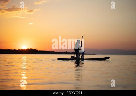 Silhouette einer Person, die bei Sonnenuntergang auf einem Paddleboard steht, mit Sonnenuntergang hinter einer Insel. Dramatischer Himmel und ruhiges Wasser. Stockfoto