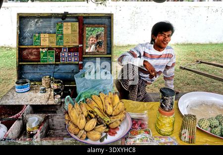 Ein Verkäufer steht auf einem Straßenmarkt in Yangon und verkauft frisches Obst und Waren an Kunden. Der Markt zeigt verschiedene Artikel auf einem belegten Tisch an. Stockfoto