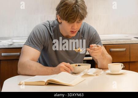Junger kaukasischer Mann, der frühstückt, während er Buch liest, in der Küche, in der Inneneinrichtung, ruhige Morgenroutine, Lifestyle und Entspannungskonzept. Morni Stockfoto