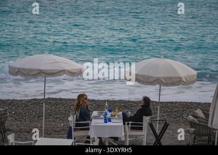 Essen am Strand in Nizza, Frankreich. Ehepaar mittleren Alters, das unter weißen Sonnenschirmen in der Nähe der stürzenden türkisfarbenen Wellen des Mittelmeers Essen genießt. Stockfoto