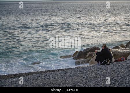 Einsame Figur, die die Wellen des Mittelmeers auf den markanten Kiesstrand von Nizza, Frankreich, schaut. Stockfoto