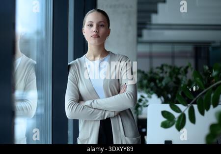 Eine Frau im Business-Anzug steht mit überkreuzten Armen vor einem Fenster Stockfoto