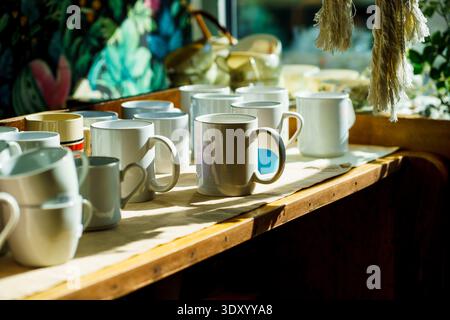 Reihe weißer Tassen im warmen Sonnenlicht auf rustikalem Regal. Café-Bar innen, morgendliche Routine und kleine Geschäftsatmosphäre. Stockfoto