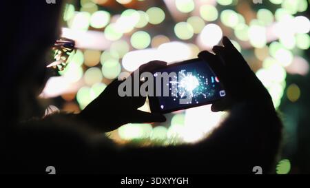 Eine Frau schießt Feuerwerk auf einem Smartphone. Die Lichter spiegeln sich wunderbar in ihrer Brille wider Stockfoto