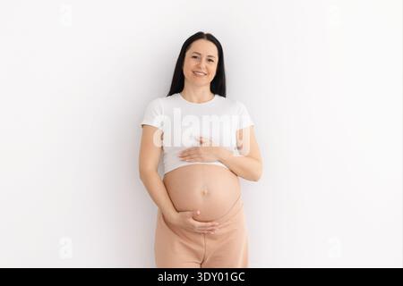 Lächelnde junge schwangere Frau zu Hause, die den Bauch berührt und in die Kamera schaut Stockfoto