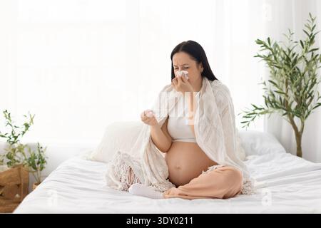 Schwangere Frau, die auf dem Bett sitzt, in eine Decke gehüllt und das Thermometer überprüft, sich unwohl fühlt Stockfoto