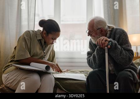 Junge Erwachsene Schwarze Sozialarbeiterin, die Dokumente mit einem älteren kaukasischen Mann überprüft Stockfoto