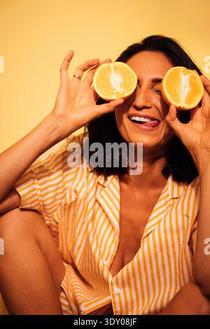 Dunkelhaarige Frau hält fröhlich orangene Hälften vor den Augen, trägt ein gestreiftes Hemd vor einem hellgelben Hintergrund, strahlt Glück und Verspieltheit aus || Model veröffentlicht Stockfoto