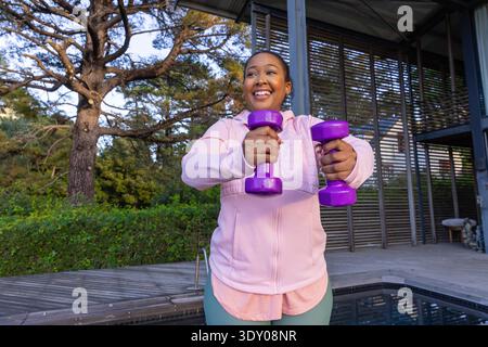 Afroamerikanische Frau, die lila Kurzhanteln nach vorne auf hölzernem Pooldeck am dunklen gefliesten Pool hebt. Fitness, Wellness, Lifestyle, Outdoor, modern, lebendig, motiviert || Model veröffentlicht Stockfoto
