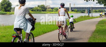 Familienradeln auf dem Radweg am Fluss nahe der Elbe in Magdeburg unter dramatischen Wolken. Mutter Kind, das Fahrrad-Helme durch grüne Landschaft reitet Stockfoto