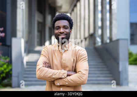 Junger Mann, der mit gekreuzten Armen steht, selbstbewusst in die Kamera lächelt und ein lässiges Langarmshirt in einer urbanen Outdoor-Umgebung in der Nähe eines modernen Gebäudes mit Stufen trägt Stockfoto