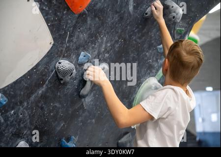 Ein Schuljunge in einem weißen T-Shirt trainiert auf einer dunklen, strukturierten Kletterwand. Er greift in einem professionellen Boulderzentrum nach grauen und blauen Holds Stockfoto