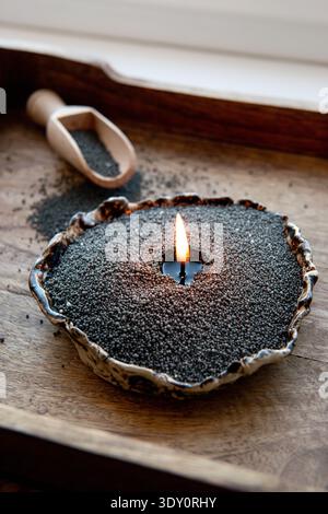 Moderne schwarze Perle Sandkerze, die in einer modernen Tonschale zu Hause brennt. Holzlöffel mit Kerzenpulver auf einem Holztablett verstreut. Stockfoto