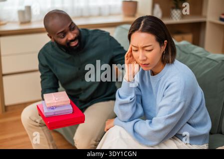 Eine enttäuschte Frau wendet sich auf dem Sofa ab, während ihr Partner einen Stapel Geschenkkartons anbietet, um einen angespannten Moment in ihrer Beziehung zu Hause festzuhalten. Stockfoto