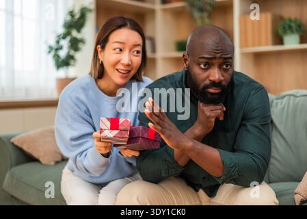 Ein Paar sitzt zusammen in einem Wohnzimmer, wo die Frau bunte Geschenkboxen hält, während der Mann besorgt oder frustriert wirkt, was auf eine Fehlinterpretation hindeutet Stockfoto